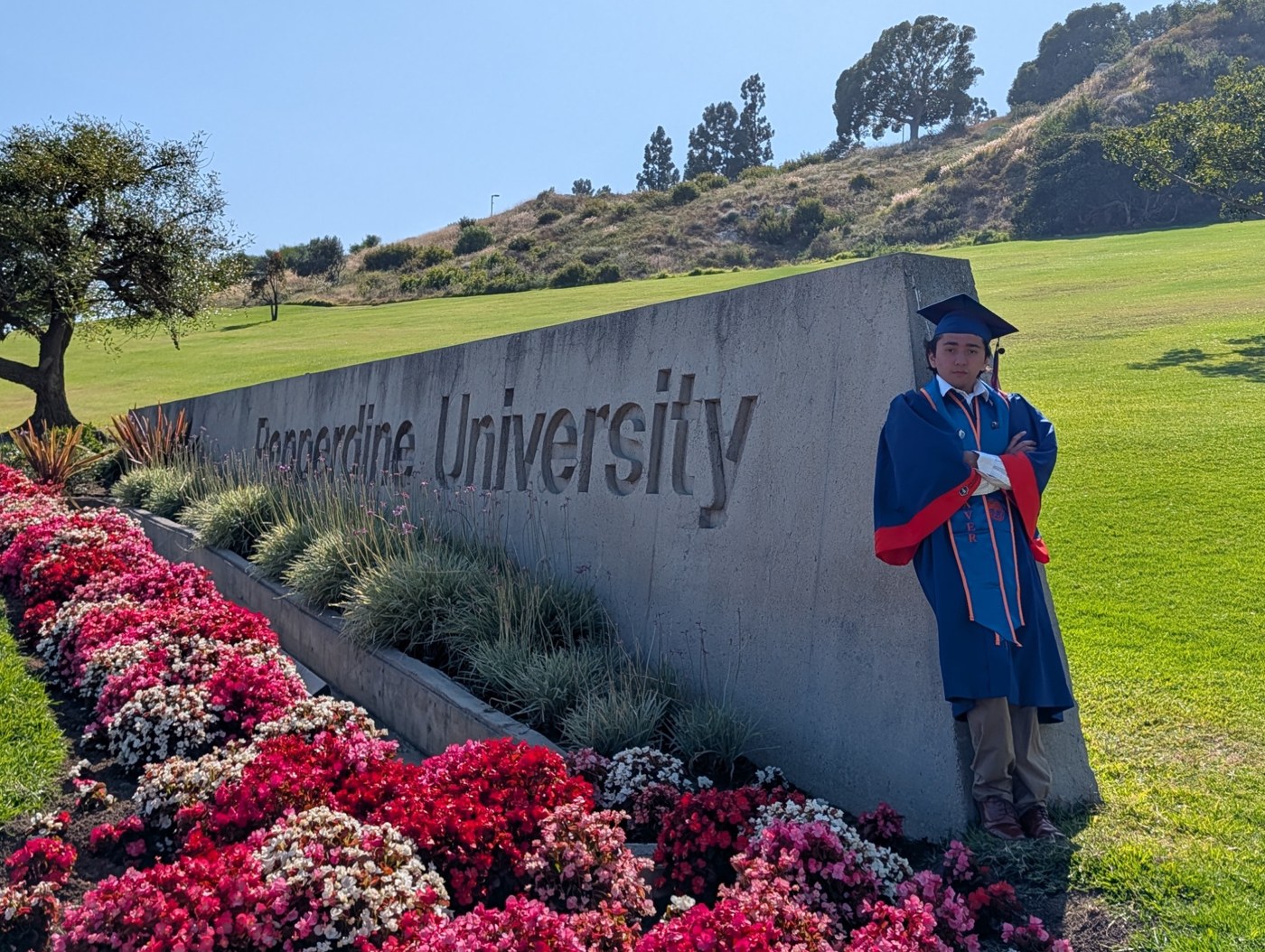 A person in graduation attire beside the Pepperdine University sign.
